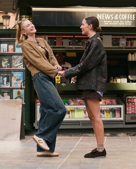 Zwei Frauen stehen lachend vor einem Kiosk und halten sich an den Händen. Sie tragen legere Outfits mit Gabor Sneakern. | © Gabor Shoes GmbH, Rosenheim