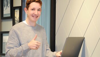 Eine Frau mit grauem Pullover steht an einem Laptop und zeigt einen Daumen nach oben.