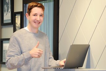 Eine Frau mit grauem Pullover steht an einem Laptop und zeigt einen Daumen nach oben.