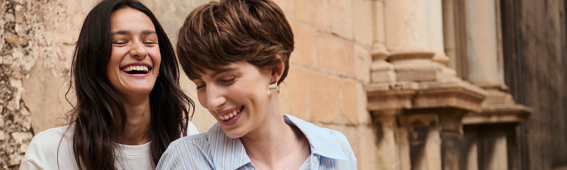 myGabor | Twee lachende vrouwen staan iets verspringend achter elkaar voor een oude stenen muur, close-up van bovenlichaam en gezichten | © Gabor Shoes GmbH, Rosenheim