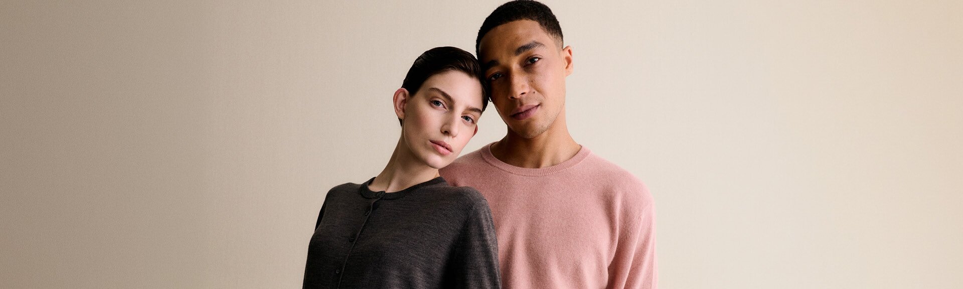 A woman in a grey sweater and a man in a pink sweater lean against each other in front of a light background and appear relaxed. | © Gabor Shoes GmbH, Germany