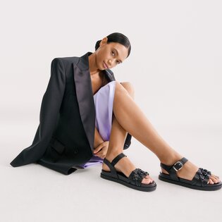 En kvinna sitter i ett ljust rum, har på sig en lila klänning och en svart kavaj samt svarta sandaler med remmar och dekorativa detaljer. | © Gabor Shoes GmbH, Tyskland