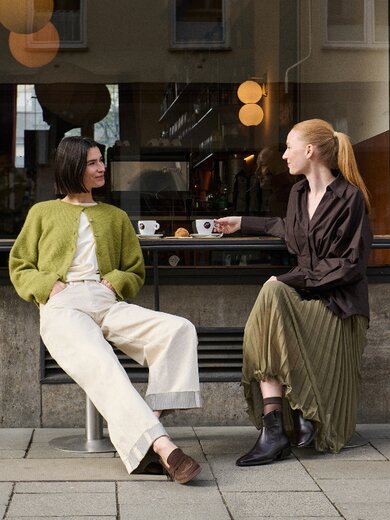 Una donna è seduta in una stanza luminosa, indossa un abito lilla e un blazer nero, e sandali neri con cinturini e dettagli decorativi. | © Gabor Shoes GmbH, Germania