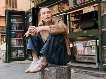 En kvinna sitter avslappnat på ett granitblock framför en öppen kiosk, håller en mugg i handen och bär ljusa Gabor-sneakers, blå jeans och en beige jacka. I bakgrunden syns drycker och magasin. | © Gabor Shoes GmbH, Tyskland