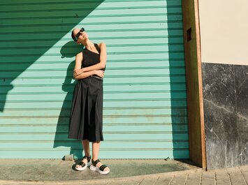 En kvinna står avslappnat med armarna i kors framför en turkos garageport. Hon bär svarta solglasögon, ett svart ärmlöst linne, svarta vida byxor och svarta sandaler med vit sula. | © Gabor Shoes GmbH, Tyskland