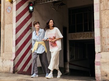 Två kvinnor går arm i arm från ett parkeringsgarage ut på trottoaren, bredvid en vägg med rödvita ränder och ett grönt trafikljus. Båda har på sig avslappnade kläder och ljusa sneakers. | © Gabor Shoes GmbH, Tyskland