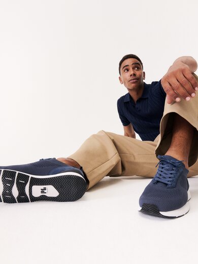 En man sitter avslappnat på golvet, har på sig beigea byxor, en blå topp och mörkblå sneakers. Skorna syns tydligt i förgrunden av bilden. | © Gabor Shoes GmbH, Tyskland