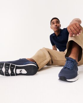 Ein Mann sitzt entspannt auf dem Boden, trägt beigefarbene Hose, blaues Oberteil und dunkelblaue Sneaker. Die Schuhe sind prominent im Vordergrund des Bildes zu sehen. | © Gabor Shoes GmbH, Rosenheim