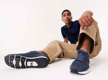 Ein Mann sitzt entspannt auf dem Boden, trägt beigefarbene Hose, blaues Oberteil und dunkelblaue Sneaker. Die Schuhe sind prominent im Vordergrund des Bildes zu sehen. | © Gabor Shoes GmbH, Rosenheim