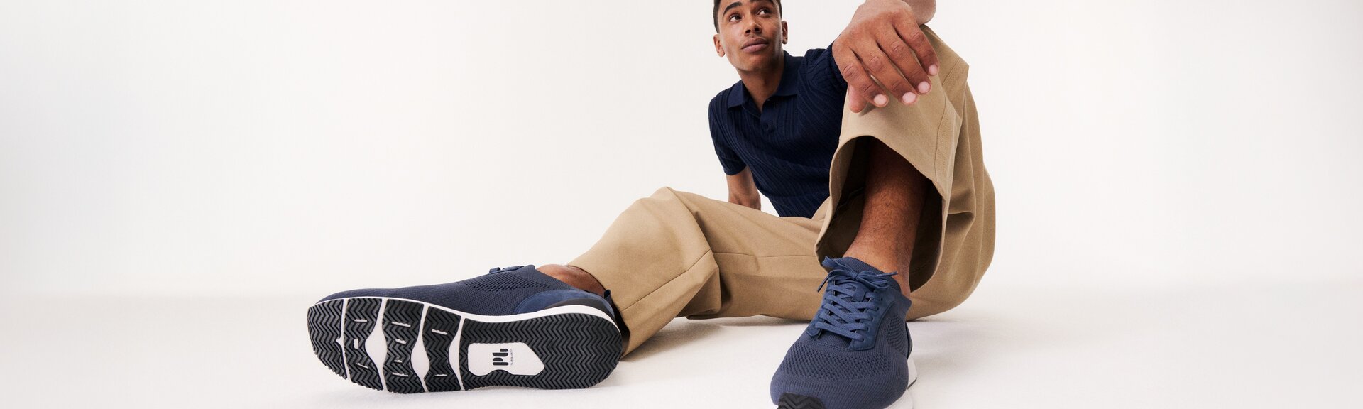 En man sitter avslappnat på golvet, har på sig beigea byxor, en blå topp och mörkblå sneakers. Skorna syns tydligt i förgrunden av bilden. | © Gabor Shoes GmbH, Tyskland