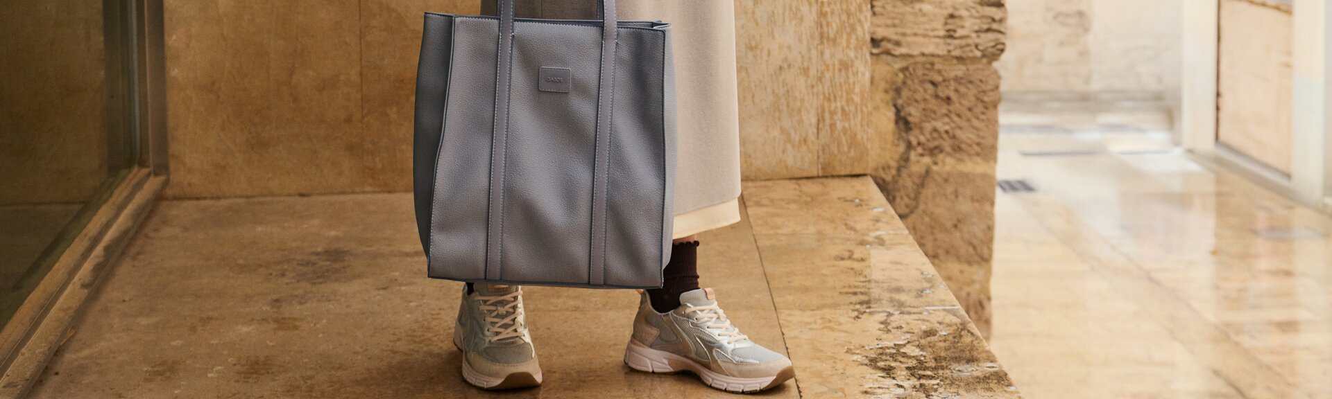 "Eine Frau steht vor einer Steinwand und trägt eine große, hellblaue Handtasche an langen Henkeln. Sie ist in einem beigen Rock, braunem Oberteil und hellen Sneakern gekleidet. " | © Gabor Shoes GmbH, Rosenheim