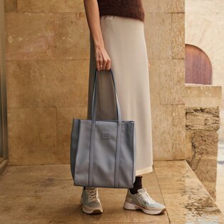 "Eine Frau steht vor einer Steinwand und trägt eine große, hellblaue Handtasche an langen Henkeln. Sie ist in einem beigen Rock, braunem Oberteil und hellen Sneakern gekleidet. " | © Gabor Shoes GmbH, Rosenheim