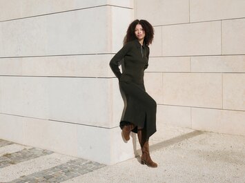 En kvinna med lockigt hår lutar mot en ljus stenmur, har på sig en lång svart klänning och bruna stövlar. Scenen är solig och lugn. | © Gabor Shoes GmbH, Tyskland