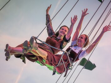 Två kvinnor sitter bredvid varandra på en kedjekarusell och sträcker glatt upp armarna. De har traditionella dirndl och bilden visar himlen i bakgrunden. | © Gabor Shoes GmbH, Tyskland