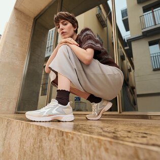 En kvinna sitter hukande på ett stensteg framför en modern byggnad och har ljusa sneakers, mörka strumpor, en vid kjol och en kortärmad tröja på sig. | © Gabor Shoes GmbH, Tyskland