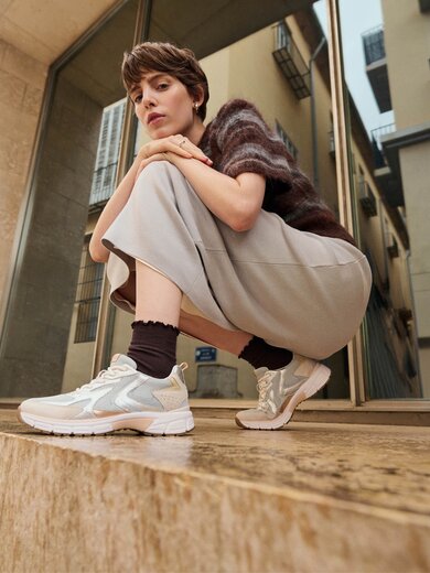 En kvinna sitter hukande på ett stensteg framför en modern byggnad och har ljusa sneakers, mörka strumpor, en vid kjol och en kortärmad tröja på sig. | © Gabor Shoes GmbH, Tyskland