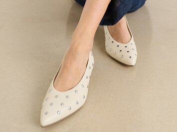 Eine Frau sitzt auf dem Boden und trägt eine dunkelblaue Hose sowie beigefarbene, spitze Pumps mit silbernen Nieten. | © Gabor Shoes GmbH, Rosenheim