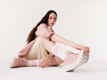 En kvinna sitter på golvet i ett ljust rum och bär rosa sneakers, randiga strumpor, en cremefärgad kjol och en rosa jacka. | © Gabor Shoes GmbH, Tyskland