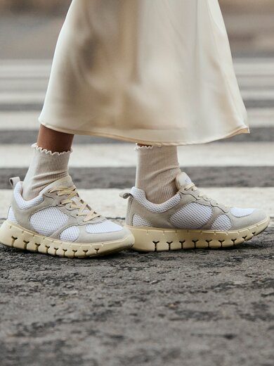 Två ben i vita sportstrumpor och krämfärgade sneakers står på ett övergångsställe. Kvinnan bär en lång, ljus kjol. | © Gabor Shoes GmbH, Tyskland