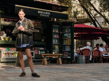 Eine Frau steht mit Kaffee vor einem Zeitschriften-Kiosk und trägt braune flache Sneaker. | © Gabor Shoes GmbH, Rosenheim