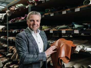 Markus Kramer, Director Purchasing bei Gabor hält ein großes Stück braunes Leder in der Hand und steht vor einem Regal mit verschiedenen Lederstücken in einer Lagerhalle. | © Gabor Shoes GmbH, Rosenheim