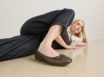 En kvinna ligger på golvet och har på sig en mörkbrun flätad ballerinasko i läder, kombinerat med mörka jeans. Fokus ligger på skon. | © Gabor Shoes GmbH, Tyskland
