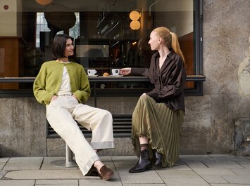 Eine Frau sitzt in einem hellen Raum, trägt ein lilafarbenes Kleid und einen schwarzen Blazer sowie schwarze Sandalen mit Riemen und dekorativen Details. | © Gabor Shoes GmbH, Rosenheim