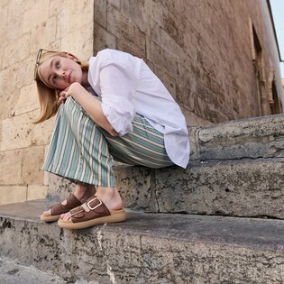 En kvinna sitter på en stentrappa framför en gammal mur. Hon har på sig en vit blus, vida randiga byxor i grönt och beige samt bruna sandaler med guldfärgat spänne. Hon har skjutit upp solglasögonen i håret. | © Gabor Shoes GmbH, Tyskland