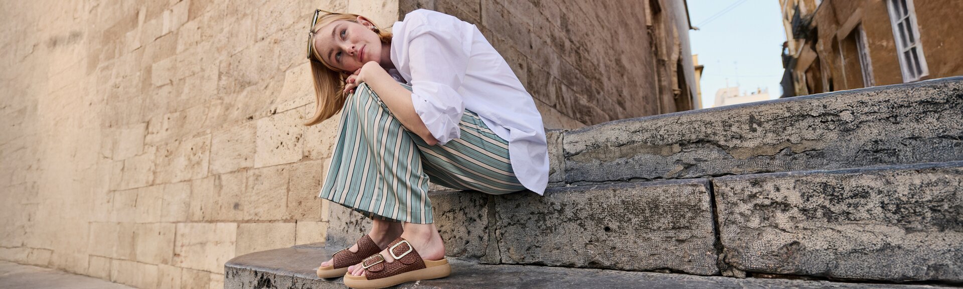 En kvinna sitter på en stentrappa framför en gammal mur. Hon har på sig en vit blus, vida randiga byxor i grönt och beige samt bruna sandaler med guldfärgat spänne. Hon har skjutit upp solglasögonen i håret. | © Gabor Shoes GmbH, Tyskland