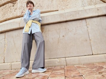 En kvinna står framför en stenmur på mönstrade golvplattor, bär en ljusblå blus, en gul stickad kofta runt höfterna, vida grå byxor och ljusblå sneakers. Bilden är tagen ur grodperspektiv. | © Gabor Shoes GmbH, Tyskland