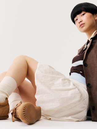 Eine Frau sitzt auf dem Boden vor weißem Hintergrund und trägt braune Sneaker, weiße Socken, einen beigefarbenen Rock, ein gestreiftes Oberteil und einen braunen Blazer. | © Gabor Shoes GmbH, Rosenheim