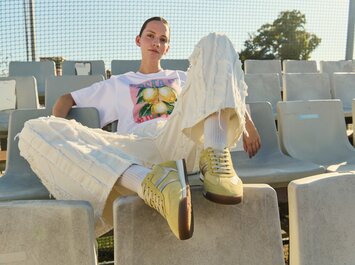 En kvinna sitter avslappnat på läktaren, har på sig ljusa, strukturerade byxor, en vit T-shirt med citronmotiv och gula sneakers. | © Gabor Shoes GmbH, Tyskland