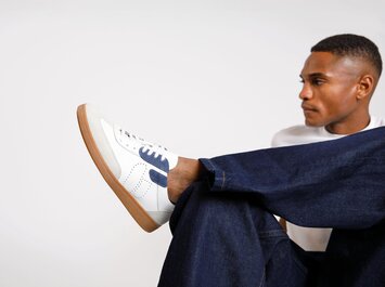 En man bär vita sneakers med blå detaljer och brun sula, tillsammans med mörkblå jeans. Skon är tydligt synlig i förgrunden. | © Gabor Shoes GmbH, Tyskland