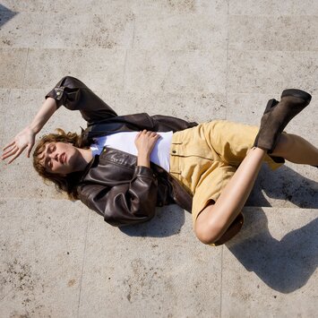 Eine Frau liegt auf steinernen Stufen. Sie trägt eine braune Lederjacke, ein weißes Oberteil, beige Shorts und braune Stiefeletten. | © Gabor Shoes GmbH, Rosenheim