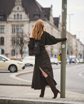 Eine Frau steht in herbstlicher Natur auf einem Kiesweg, trägt braune Stiefel, eine braune Jacke und einen kurzen Rock, und lehnt sich an trockenes, hohes Schilfgras. | © Gabor Shoes GmbH, Rosenheim