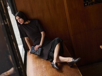 Une femme aux cheveux bouclés s’appuie contre un mur de pierre clair, porte une longue robe noire et des bottes marron. La scène est ensoleillée et calme. | © Gabor Shoes GmbH, Allemagne