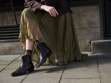 Eine Frau trägt dunkelbraune Damen-Stiefeletten mit spitzer Kappe und kleinem Absatz zu einem langen, fließenden olivgrünen Rock; sie steht auf einem gepflasterten Gehweg vor Steinstufen. | © Gabor Shoes GmbH, Rosenheim