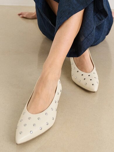 Una donna è seduta a terra e indossa pantaloni blu scuro e décolleté beige a punta con borchie argentate. | © Gabor Shoes GmbH, Germania