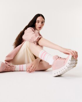 Eine Frau sitzt auf dem Boden in einem hellen Raum und trägt rosa Sneaker, gestreifte Socken, einen cremefarbenen Rock und eine rosa Jacke. | © Gabor Shoes GmbH, Rosenheim