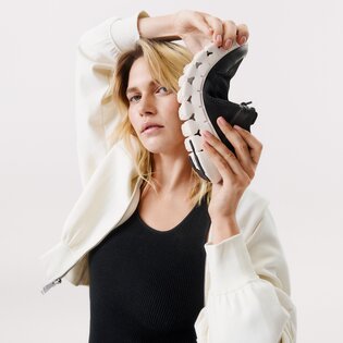 En kvinna håller en flexibel svart sneaker med vit sula bredvid sitt huvud. Hon har på sig en vit jacka och svart topp mot ljus bakgrund. | © Gabor Shoes GmbH, Tyskland