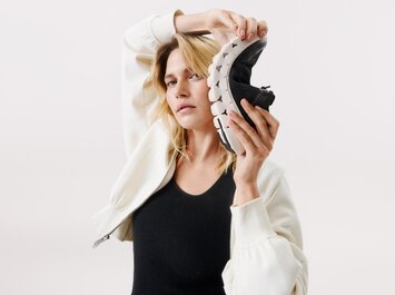 En kvinna håller en flexibel svart sneaker med vit sula bredvid sitt huvud. Hon har på sig en vit jacka och svart topp mot ljus bakgrund. | © Gabor Shoes GmbH, Tyskland