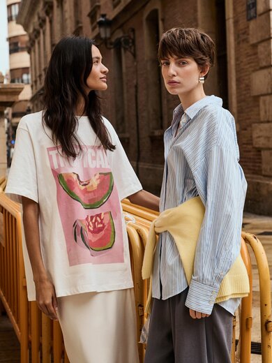 Två kvinnor står bredvid varandra på en våt gata framför en gammal byggnad; den ena bär en vit t-shirt med vattenmelontryck och texten ”Tropical”, den andra har på sig en ljusblå skjorta och en gul tröja knuten runt höfterna. I bakgrunden står cyklar lutade mot ett räcke. | © Gabor Shoes GmbH, Tyskland