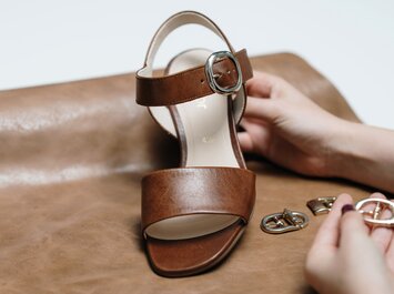 Eine braune Sandale mit silberner Schnalle wird auf braunem Leder präsentiert. Eine Hand hält die Sandale, daneben liegen weitere Schnallen. | © Gabor Shoes GmbH, Rosenheim