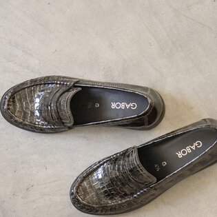 Eine Frau  sitzt entspannt auf dem Boden in einem hellgelben Blazer mit Gürtel und passender Shorts. Sie trägt helle Sandalen mit Absatz, die Beine sind seitlich ausgestreckt. Der Hintergrund ist neutral und schlicht. | © Gabor Shoes GmbH, Rosenheim