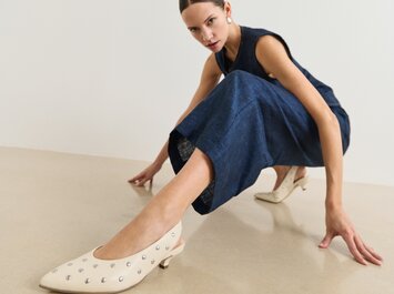 En kvinna i mörkblå ärmlös jumpsuit visar upp cremefärgade skor med små silvernitar och låg klack på ett ljust golv mot vit bakgrund. | © Gabor Shoes GmbH, Tyskland