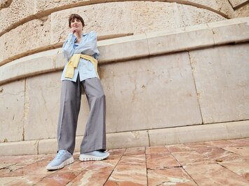 En kvinna står framför en ljus stenmur och har på sig gråa vida byxor, en ljusblå skjorta och en gul tröja knuten runt midjan. Ljusa blå sneakers syns på ett mönstrat stengolv. | © Gabor Shoes GmbH, Tyskland