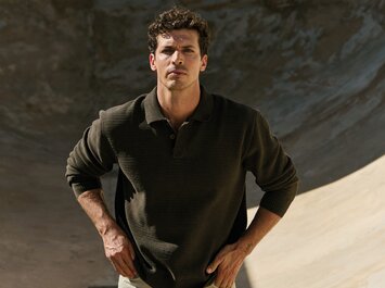 En man står med händerna i sidan framför en rund, betonbakgrund och bär en mörkgrön stickad tröja med krage och beige byxor. | © Gabor Shoes GmbH, Tyskland