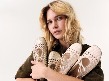 Eine blonde Frau hält mehrere beige und goldene, geflochtene Ballerinas mit Lochmuster im Arm, vor hellem Hintergrund. | © Gabor Shoes GmbH, Rosenheim