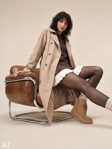 Une image générée par l’IA montrant une femme assise dans un fauteuil en cuir devant un fond beige. Elle porte un trench-coat brun clair sur un haut brun foncé et une jupe plissée blanche, et elle a des bottines à lacets beiges de Gabor aux pieds. | © Gabor Shoes GmbH, Allemagne