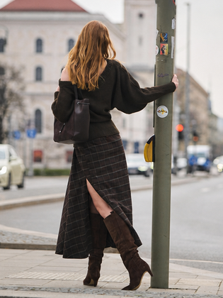 Eine Frau steht in herbstlicher Natur auf einem Kiesweg, trägt braune Stiefel, eine braune Jacke und einen kurzen Rock, und lehnt sich an trockenes, hohes Schilfgras. | © Gabor Shoes GmbH, Rosenheim